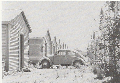 A black and white photo showing a car parked on a driveway of a woodern hut 