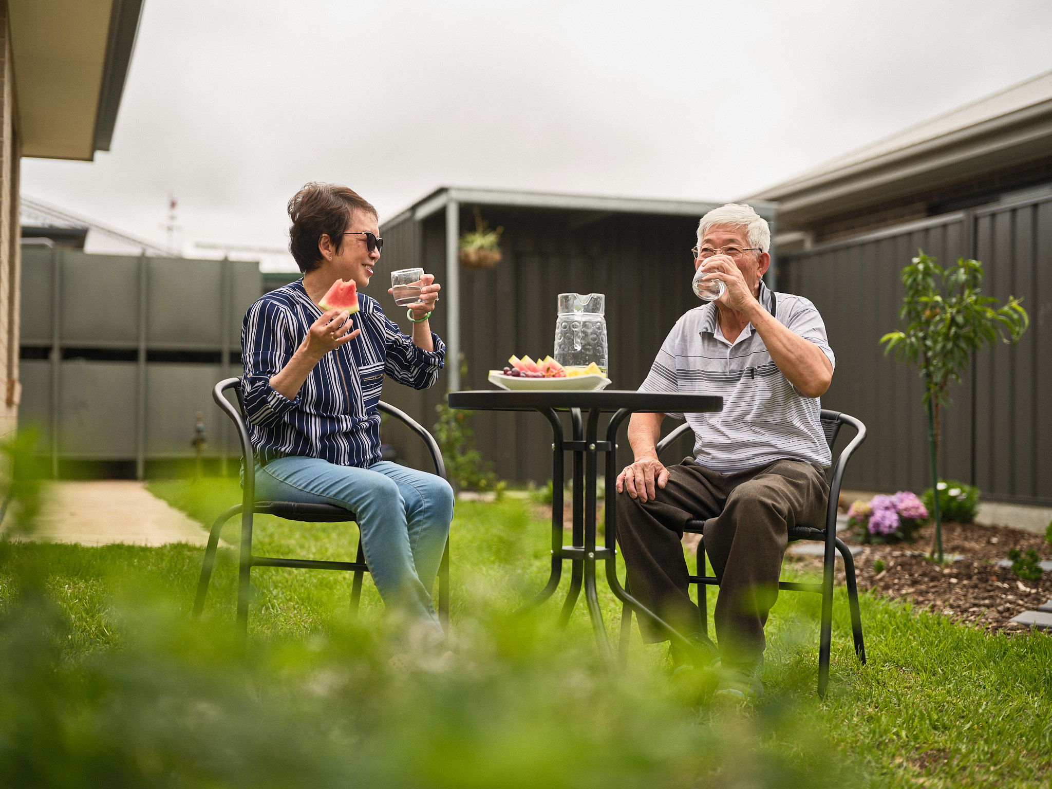 An older adult man with glasses pours water from a clear pitcher into a glass at an outdoor seating area, engaging in a casual conversation with a lady drinking water and eating watermelon. The setting suggests a warm, friendly atmosphere with a focus on hydration and the importance of clean drinking water.